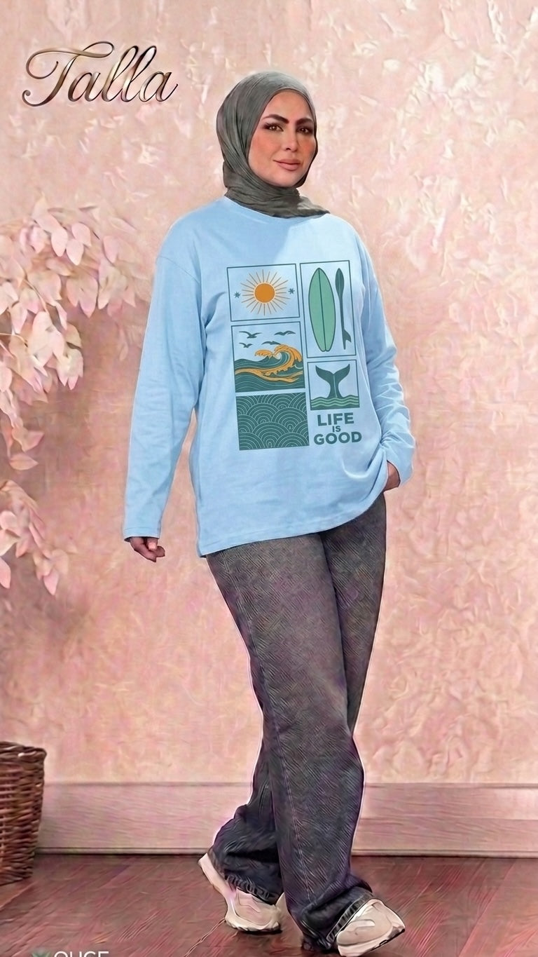 Model wearing a Talla | طلةّ light blue long-sleeve graphic sweatshirt with sun and surf motifs and grey trousers.