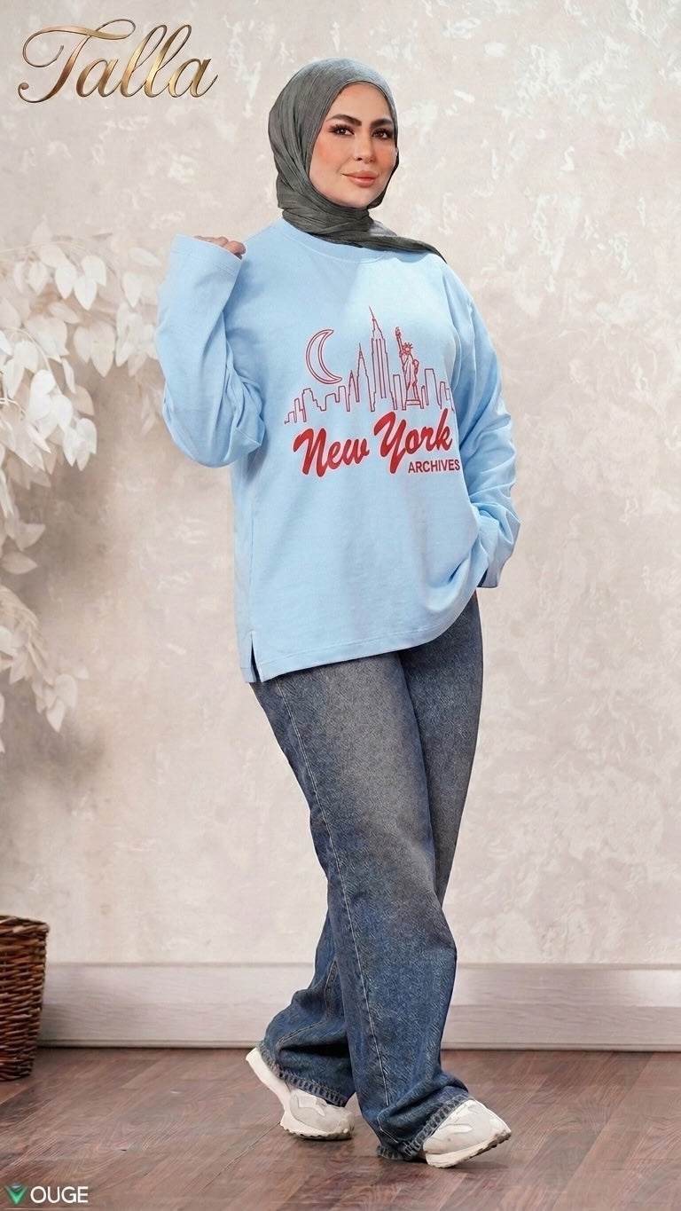 Talla | طلةّ light blue crewneck sweatshirt with New York skyline graphic, worn by a woman in a grey hijab and jeans.