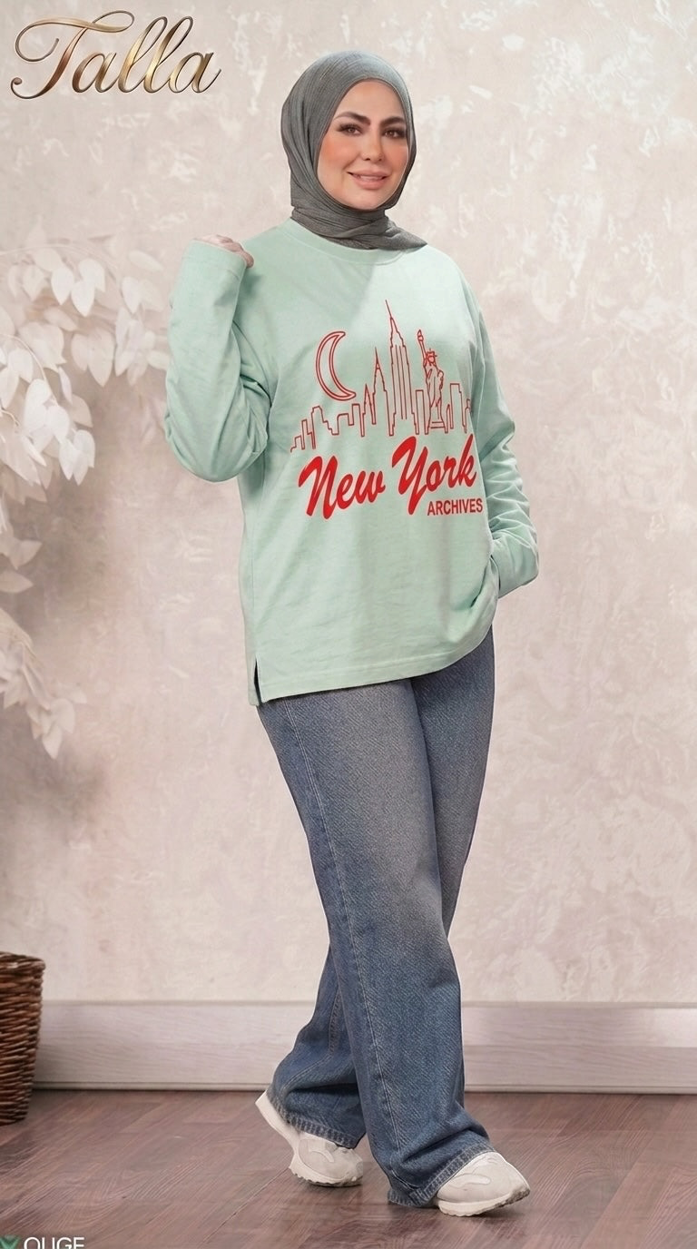 Talla | طلةّ mint green crewneck sweatshirt with New York skyline graphic, worn by a woman in a grey hijab and jeans.